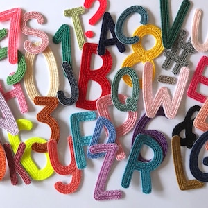 Könnte beinhalten: Eine Sammlung bunter, gestrickter Buchstaben und Zahlen, die auf einer weißen Oberfläche verstreut sind. Die Buchstaben und Zahlen sind in verschiedenen Farben gehalten, darunter Rosa, Blau, Grün, Gelb und Rot. Das Bild ist gut beleuchtet und die Details der Strickstruktur sind sichtbar.