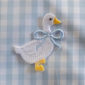 Peut inclure: Une broderie blanche représentant une oie avec un nœud bleu clair et des pieds jaunes est placée sur un tissu à carreaux vichy bleu clair et blanc. Le motif de l'oie est charmant et fantaisiste, idéal pour les articles de bébé ou la décoration de chambre d'enfant.
