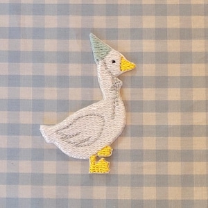 Op de afbeelding: Witte geborduurde gans met een lichtblauwe feestmuts met gele accenten. De gans staat op een lichtblauwe en witte ruitjesachtergrond.