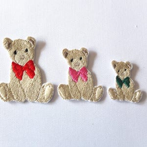 Può includere: Tre toppe ricamate a forma di orsetto di diverse dimensioni. Ogni orsetto è beige con un fiocco di colore diverso: rosso, rosa e verde. Le toppe sono su una superficie bianca.