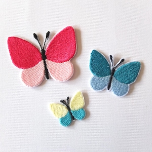 Op de afbeelding: Drie geborduurde strijkpatches in de vorm van vlinders. De grootste vlinder is roze en rood. De middelste vlinder is blauw en turquoise. De kleinste vlinder is geel en turquoise.
