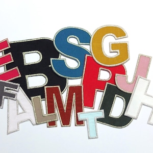 May include: A selection of fabric letters in a variety of colours, such as red, black, blue, and yellow. Each letter has a silver trim. The letters are arranged on a white background.