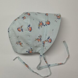 Puede incluir: Un gorro azul claro con estampado floral y lunares blancos. El gorro tiene dos lazos largos que se pueden atar debajo de la barbilla.