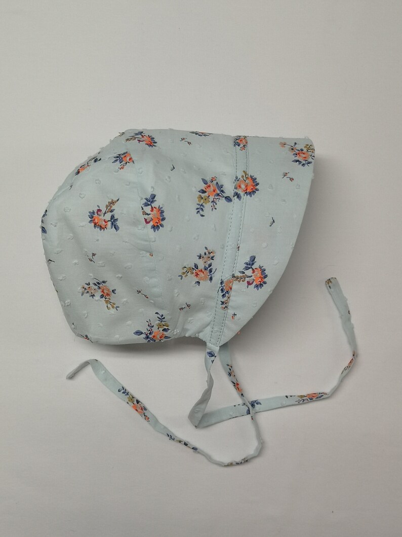 Puede incluir: Un gorro azul claro con estampado floral y lunares blancos. El gorro tiene dos lazos largos que se pueden atar debajo de la barbilla.