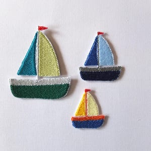 Op de afbeelding: Drie geborduurde vilt zeilboot patches in verschillende maten. De grootste zeilboot is groen, geel en blauw. De middelste zeilboot is blauw en wit. De kleinste zeilboot is blauw, geel en oranje.