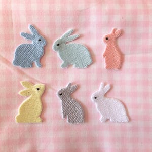 Op de afbeelding: Zes geborduurde konijnenpatches in verschillende pastelkleuren, waaronder lichtblauw, lichtroze, lavendel en geel. De patches bevinden zich op een roze-witte ruitjesachtergrond.