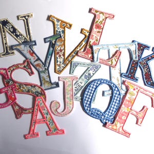 Peut inclure: Diverses lettres en tissu aux motifs et couleurs variés, notamment floraux et à carreaux. Chaque lettre est bordée d'une couleur contrastante. Les lettres sont disposées sur un fond blanc.