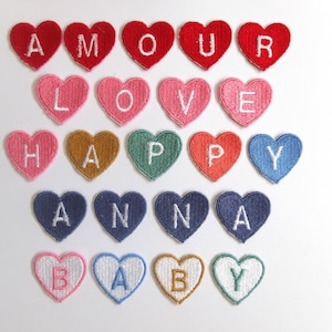 May include: A collection of heart-shaped patches in various colors, each embroidered with a single letter. The patches spell out words like "AMOUR", "LOVE", "HAPPY", "ANNA", and "BABY". The patches are arranged on a white background.