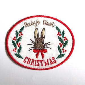 Könnte beinhalten: Gestickter Aufnäher mit einem weißen ovalen Mittelteil und einem roten Rand. Der Aufnäher zeigt ein braunes Häschen mit roter Schleife, umgeben von dem Text "Baby's First Christmas". Grüne Blätter und rote Beeren umrahmen das Häschen.