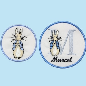 Può includere: Due toppe ricamate con un coniglio dei cartoni animati con una giacca blu. Una toppa è un semplice cerchio con il coniglio, l'altra ha la lettera "M" e il nome "Marcel". Entrambe le toppe hanno un bordo blu e sono su sfondo bianco.