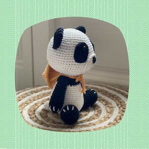 May include: A crocheted panda plush toy with black and white fur, wearing a yellow and orange scarf. The panda is sitting on a woven rug.