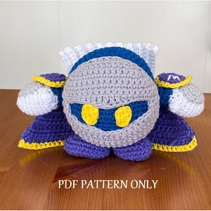May include: Crocheted plush toy of a gray and blue character with yellow eyes and a purple cape. The toy is sitting on a wooden surface. PDF PATTERN ONLY.