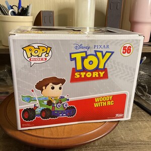 Op de afbeelding: Een Funko Pop! Rides doos met Woody uit Toy Story in een groene en blauwe RC auto. De doos heeft het Toy Story logo, nummer 56 en de tekst "Woody with RC".
