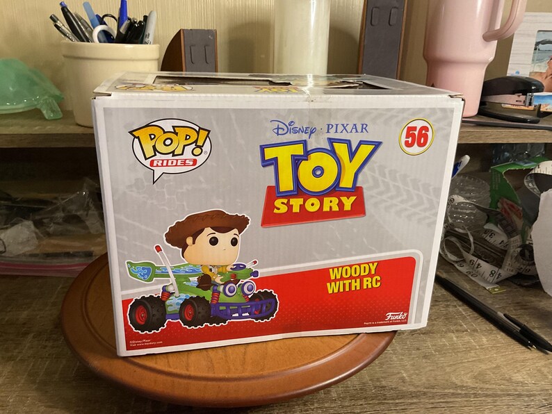 Op de afbeelding: Een Funko Pop! Rides doos met Woody uit Toy Story in een groene en blauwe RC auto. De doos heeft het Toy Story logo, nummer 56 en de tekst "Woody with RC".