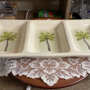 May include: A rectangular, off-white serving dish with three compartments. Each section features a hand-painted palm tree design in green and brown. The dish is resting on a round, brown table covered with a white lace doily.