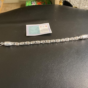 May include: A silver bracelet with an oval link design and a magnetic clasp. The bracelet is displayed on a black surface, with a small white card nearby. The bracelet's links have a textured pattern.