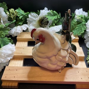 May include: A white ceramic chicken figurine with green and black painted details. The chicken is sitting on a wooden pallet with white flowers in the background.