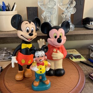 May include: Three Mickey Mouse figurines of varying sizes stand on a wooden surface. The largest Mickey wears a black body, red shorts with yellow buttons, and a yellow bow tie. The middle Mickey wears a red shirt. The smallest Mickey is in a yellow shirt and blue pants.