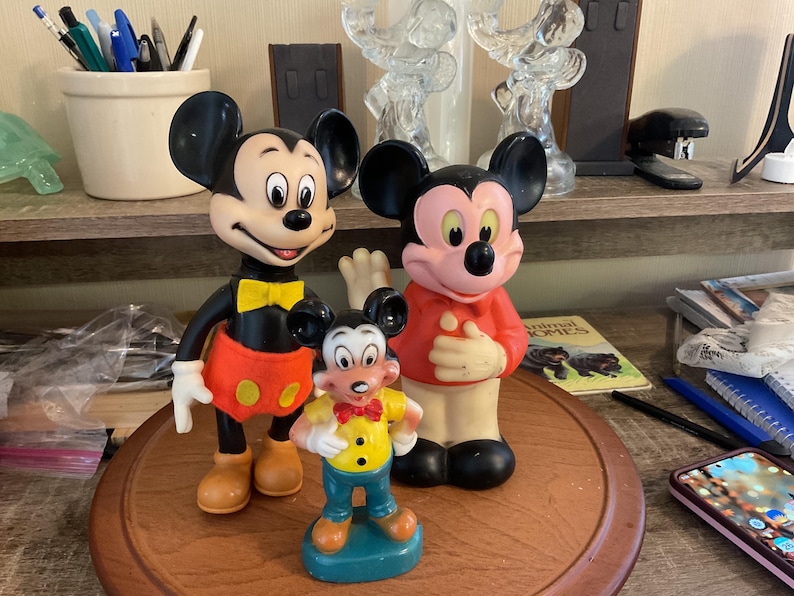 May include: Three Mickey Mouse figurines of varying sizes stand on a wooden surface. The largest Mickey wears a black body, red shorts with yellow buttons, and a yellow bow tie. The middle Mickey wears a red shirt. The smallest Mickey is in a yellow shirt and blue pants.