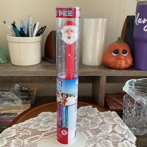May include: A red PEZ dispenser featuring a Santa Claus head and body, encased in a clear plastic tube. The dispenser has a blue and white label with a winter scene. The dispenser is on a wooden shelf.