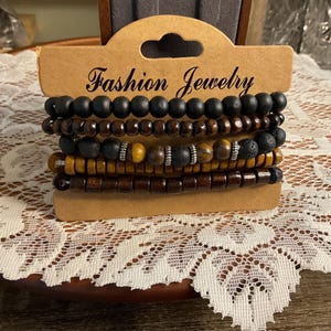May include: A set of five bracelets with various beads, including black, brown, and yellow tones. The bracelets are displayed on a brown cardboard tag that reads "Fashion Jewelry". The bracelets are on a white lace surface.