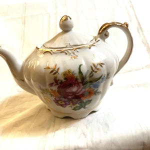 May include: White porcelain teapot with a floral design and gold accents. The teapot has a round body and a lid with a knob.