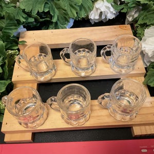 Puede incluir: Seis vasos de chupito de vidrio transparente con forma de jarra de cerveza dispuestos en dos filas sobre una superficie de madera. Los vasos están vacíos y tienen una superficie texturizada.