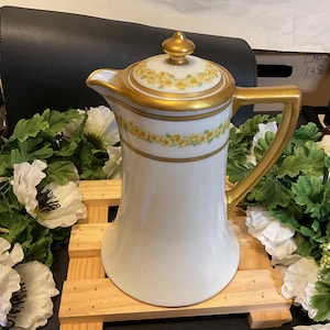 May include: White porcelain coffee pot with a gold trim and yellow floral design. The pot has a lid with a gold knob and a handle.