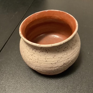 May include: A small, white ceramic pot with a speckled finish and a reddish-brown interior. The pot is round and has a wide opening.