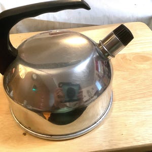 May include: A silver kettle with a black handle and a black knob on the spout. The kettle is sitting on a wooden surface.