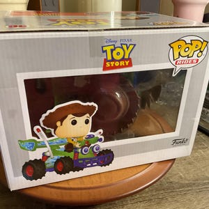 Op de afbeelding: Een Funko Pop! Rides doos met Woody uit Disney Pixar's Toy Story. De doos heeft een helder venster met de figuur van Woody in een groene en paarse raceauto. De doos heeft de Toy Story en Pop! Rides logo's.