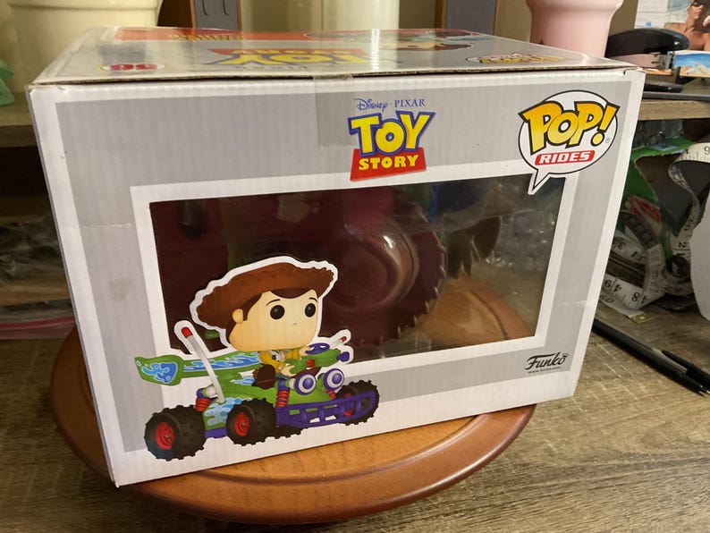 Op de afbeelding: Een Funko Pop! Rides doos met Woody uit Disney Pixar's Toy Story. De doos heeft een helder venster met de figuur van Woody in een groene en paarse raceauto. De doos heeft de Toy Story en Pop! Rides logo's.