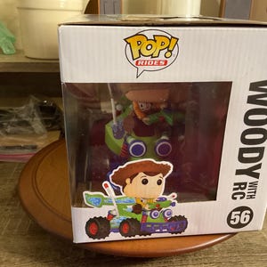 Op de afbeelding: Een Funko Pop! Rides figuur met Woody uit Toy Story in een groene en blauwe RC-auto. De witte doos heeft de tekst "Woody with RC" en het nummer 56. Het figuur zit in een doorzichtige plastic display case.