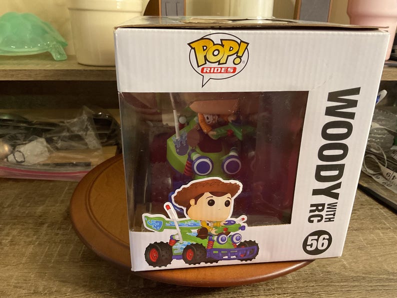 Op de afbeelding: Een Funko Pop! Rides figuur met Woody uit Toy Story in een groene en blauwe RC-auto. De witte doos heeft de tekst "Woody with RC" en het nummer 56. Het figuur zit in een doorzichtige plastic display case.