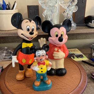 May include: Three Mickey Mouse figurines. The largest Mickey is black, red, and yellow. The middle Mickey is red and pink. The smallest Mickey is yellow, blue, and orange. All three are standing on a wooden surface.