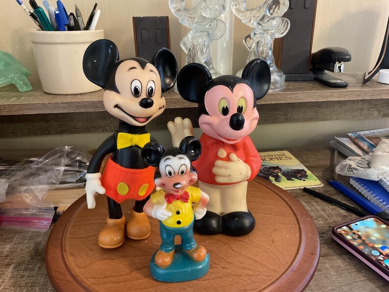 May include: Three Mickey Mouse figurines. The largest Mickey is black, red, and yellow. The middle Mickey is red and pink. The smallest Mickey is yellow, blue, and orange. All three are standing on a wooden surface.