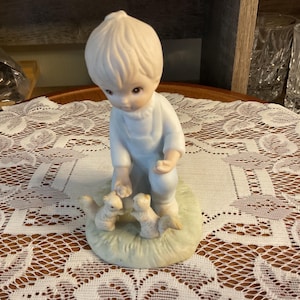 May include: A porcelain figurine of a young child in a light blue outfit, kneeling on a patch of green grass. The child is interacting with three small squirrels. The figurine has a soft, pastel color palette.
