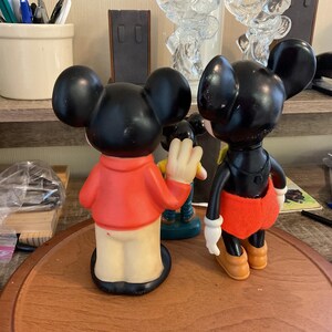 May include: Two vintage Mickey Mouse figurines. One Mickey is wearing a red shirt and tan pants, the other is wearing a black shirt, red shorts, and yellow shoes. Both have black ears and are standing on a wooden surface.