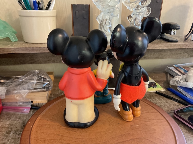 May include: Two vintage Mickey Mouse figurines. One Mickey is wearing a red shirt and tan pants, the other is wearing a black shirt, red shorts, and yellow shoes. Both have black ears and are standing on a wooden surface.