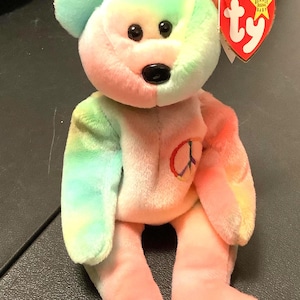 May include: A tie-dye plush bear with a peace sign on its chest. The bear is sitting with its legs crossed and has a red Ty tag attached to its ear. The tag says "Beanie Babies Original Baby".