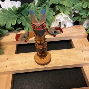 May include: A small wooden totem pole with a carved bird figure at the top. The bird is painted in blue, red, yellow, and black. The totem pole is standing on a wooden base.