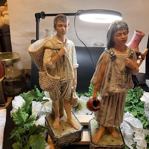 Può includere: Due figurine in ceramica di persone, una con un cesto e l'altra con una brocca. Entrambe le figurine sono in piedi su una base con un design decorativo.