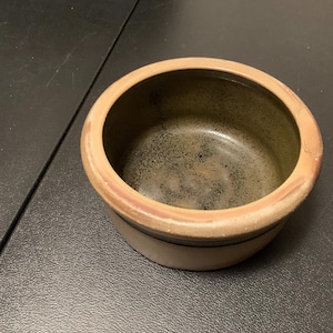May include: A small, round, ceramic bowl with a brown and black glaze. The bowl has a slightly raised rim and a smooth, rounded bottom.