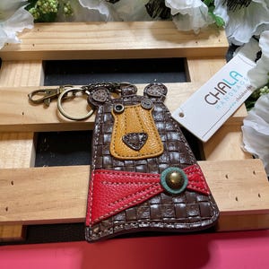 May include: A brown leather keychain with a bear face design. The bear has a yellow nose and red and brown accents. The keychain is attached to a silver key ring. The keychain is labeled "CHALA HANDBAGS".
