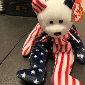 May include: A white Beanie Baby bear wearing a red, white, and blue striped outfit with a blue and white star pattern on the arms and legs. The bear has a red Ty tag attached to its ear.