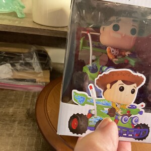 Op de afbeelding: Een Funko Pop! figuur van Woody uit Toy Story in een doorzichtige displaydoos. Woody wordt afgebeeld in een go-kart met een groen en blauw ontwerp. Het figuur zit in een doorzichtige plastic display.