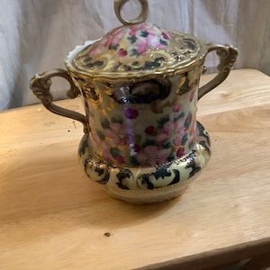 May include: A vintage porcelain sugar bowl with a lid. The bowl is decorated with pink and gold floral patterns and black accents.