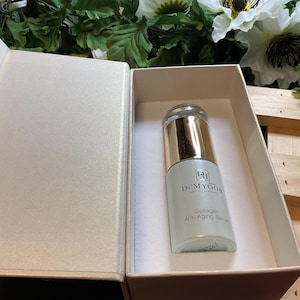 May include: A light blue serum bottle with gold accents is displayed inside a cream-colored box. The bottle is labeled "D'Myoo Collagen Anti-Aging Serum". The box is open, revealing the product, and is set against a backdrop of white flowers and greenery.