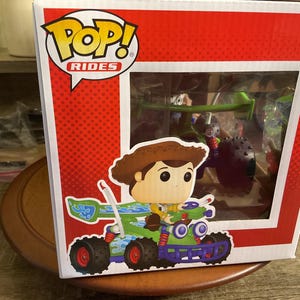 Op de afbeelding: Een Funko Pop! Rides doos met een Woody-figuur op een groen en blauw voertuig met rode wielen. De doos heeft een rode achtergrond met witte stippen en een helder venster met andere personages.