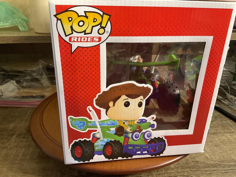 Op de afbeelding: Een Funko Pop! Rides doos met een Woody-figuur op een groen en blauw voertuig met rode wielen. De doos heeft een rode achtergrond met witte stippen en een helder venster met andere personages.